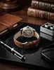 A watch and accessories are laid out on a dark surface, with books and a notebook in the background. Náhled obrázku produktu