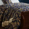 A close-up view of a navy blue and gold patterned throw blanket with fringe, draped over the side of a wooden sleigh bed. Produktbild-Vorschau