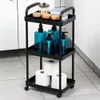 A black three-tier rolling cart filled with bathroom products like soap dispensers and toilet paper, placed on a tiled floor next to a white cabinet. Voir les images du produit