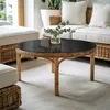 A round coffee table with a black top and rattan base, placed in a living room setting with sofas and cushions. Podgląd zdjęcia produktu