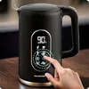 A hand is shown interacting with the digital display of a black electric kettle with gold accents, which is sitting on a wooden surface. Steam is rising from the kettle. Náhled obrázku produktu