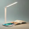 A white LED desk lamp is shown with a smartphone charging on its base, next to an open book. Produktbild-Vorschau