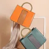 Two handbags are showcased, one orange and one teal, against a soft, peachy background with a white framed element and draped fabric. Produktbild-Vorschau