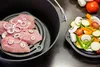 A cast iron pot with a rack holding raw meat and sliced vegetables on a dark surface. Produktbild-Vorschau