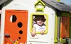 A young child wearing a hat peeks out of the window of a colorful playhouse. Produktbild-Vorschau
