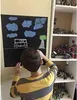 A young boy is drawing on a blackboard with a car, clouds, and a tree. There are shelves filled with toys in the background. Anteprima dell’immagine prodotto