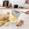 A person is using a potato ricer to press cooked potatoes into a bowl, with raw potatoes, cooked potato cakes, and a tomato on a wooden board and table. Anteprima dell’immagine prodotto