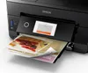 A black Epson printer is printing a document with an image of a sandwich on it. The printer's screen shows a progress indicator. Náhled obrázku produktu