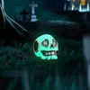 A glowing green skull charm with heart-shaped eye sockets sits on the grass in a graveyard at night. Produktbild-Vorschau