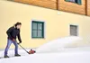 A man is using a snow blower to clear snow in front of a yellow building. Produktbild-Vorschau