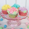 A close-up shot of colorful cupcakes decorated with frosting flowers, arranged on a pink glass cake stand, with scattered candy eggs around the base. Anteprima dell’immagine prodotto