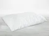 A white quilted pillow on a light gray surface with a white background. Produktbild-Vorschau
