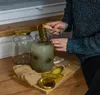 A person is placing a pickle into a jar of brine, with bread and another jar nearby. Podgląd zdjęcia produktu