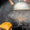 A close-up shot of a burger being cooked on a grill, with a metal dome being placed over it, creating steam. Anteprima dell’immagine prodotto