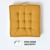A yellow square cushion with tufted details and a handle on top, set against a white background. A circular graphic with a hand sewing and text is present in the bottom left corner. Náhled obrázku produktu