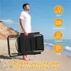 A man holding a folded black lounge chair on a sandy beach with the ocean in the background. Produktbild-Vorschau