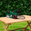 A cordless chainsaw is placed on a wooden table outdoors, with a green hedge in the background. Produktbild-Vorschau