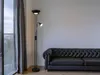A black floor lamp with a reading light is positioned next to a black tufted sofa in a room with a large window and grey curtains. Produktbild-Vorschau