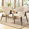 Two beige dining chairs with black legs are placed on a rug in front of a wooden dining table, with a vase of greenery on the table and a window in the background. Produktbild-Vorschau