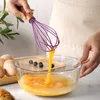 A person is whisking eggs in a glass bowl with a purple silicone whisk with a wooden handle. Produktbild-Vorschau