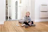 A baby sits on a wooden floor in front of a white metal baby gate, playing with toys. Produktbild-Vorschau