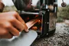 A person is adding wood to a rocket stove outdoors. Produktbild-Vorschau