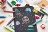 A collection of art supplies, including colored pencils and crayons, are arranged around a black sheet of paper with drawings of jellyfish. Voir les images du produit