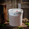 A white plastic fermentation bucket with a lid, airlock, and handle, placed on a wooden surface with moss and driftwood. The background is a rustic wooden door. Anteprima dell’immagine prodotto