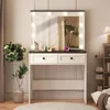 A white and black vanity table with a large mirror and lights, set up in a room with plants and a bookshelf. Voir les images du produit