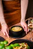 A person's hands are placing a bowl of soup on a table next to a glass of white wine and another plate of food. Náhľad obrázku produktu
