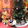 Two young children are playing with Christmas decorations around a Christmas tree and a large teddy bear in front of a fireplace backdrop. Produktbild-Vorschau