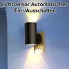 A black cylindrical light fixture plugged into a wall outlet, emitting warm light. Produktbild-Vorschau