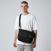 A man wearing a white t-shirt and grey pants stands against a white background, wearing a black Eastpak Delegate Plus messenger bag across his body. Produktbild-Vorschau