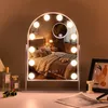 A vanity mirror with lights is placed on a desk in front of a bedroom scene reflected in the mirror. There are also perfume bottles and a framed picture on the desk. Produktbild-Vorschau