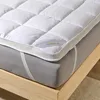 A close-up view of a white mattress topper placed on a gray mattress, with a wooden bed frame visible. Produktbild-Vorschau