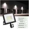 A black LED floodlight with a motion sensor is shown in the foreground, with a dark building with loading docks illuminated by other floodlights in the background. Produktbild-Vorschau