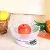 A digital kitchen scale with a clear bowl on top, holding a red apple. To the left, a basket with a yellow fruit, and to the right, a pineapple. The background is a neutral kitchen setting. Voir les images du produit