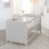 A baby crib in a nursery setting with artwork on the wall and sheer curtains. Produktbild-Vorschau