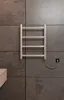 A silver electric towel warmer is mounted on a dark gray tiled wall in a bathroom. A power cord is visible, connected to a black electrical outlet. Produktbild-Vorschau