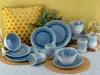 A collection of blue and white ceramic dishes, including plates, bowls, and cups, arranged on a textured surface with a yellow honeycomb-patterned pillow in the background. Náhľad obrázku produktu
