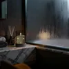 A candle with fairy lights in a glass holder sits on a marble countertop next to a diffuser and towels, with a steamy window in the background. Produktbild-Vorschau