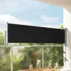 A black retractable side awning is installed on a balcony railing, providing privacy and shade. The awning is extended, and the background shows greenery and a blue sky with clouds. Anteprima dell’immagine prodotto