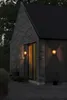 Exterior view of a modern house at dusk, with two wall lights illuminating the entrance. Produktbild-Vorschau