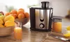 A juicer is shown on a countertop with bowls of fruit and a glass of juice. Náhľad obrázku produktu