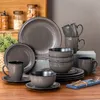 A collection of dark gray ceramic dinnerware, including plates, bowls, and mugs, arranged on a wooden table. Produktbild-Vorschau