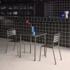 A table and two chairs set up in what appears to be an indoor cafe or restaurant setting with a black tiled wall. Produktbild-Vorschau