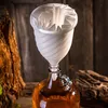 A white funnel with pleated filter paper is placed on top of a glass bottle filled with amber liquid. The background is rustic, with wooden elements and textures. Anteprima dell’immagine prodotto