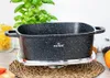 A speckled black roasting pan sits on a silver trivet, with a woven mat underneath. In the background, a potted basil plant and jars of pasta and spices are visible. Produktbild-Vorschau
