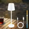 A white cordless LED table lamp is placed on a wooden table outdoors, next to a glass of dark liquid. A USB charging cable and a brightness slider are also visible. Náhľad obrázku produktu