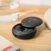A pair of black wireless earbuds in an open charging case, placed on a wooden surface next to a glass of water and a book. Produktbild-Vorschau